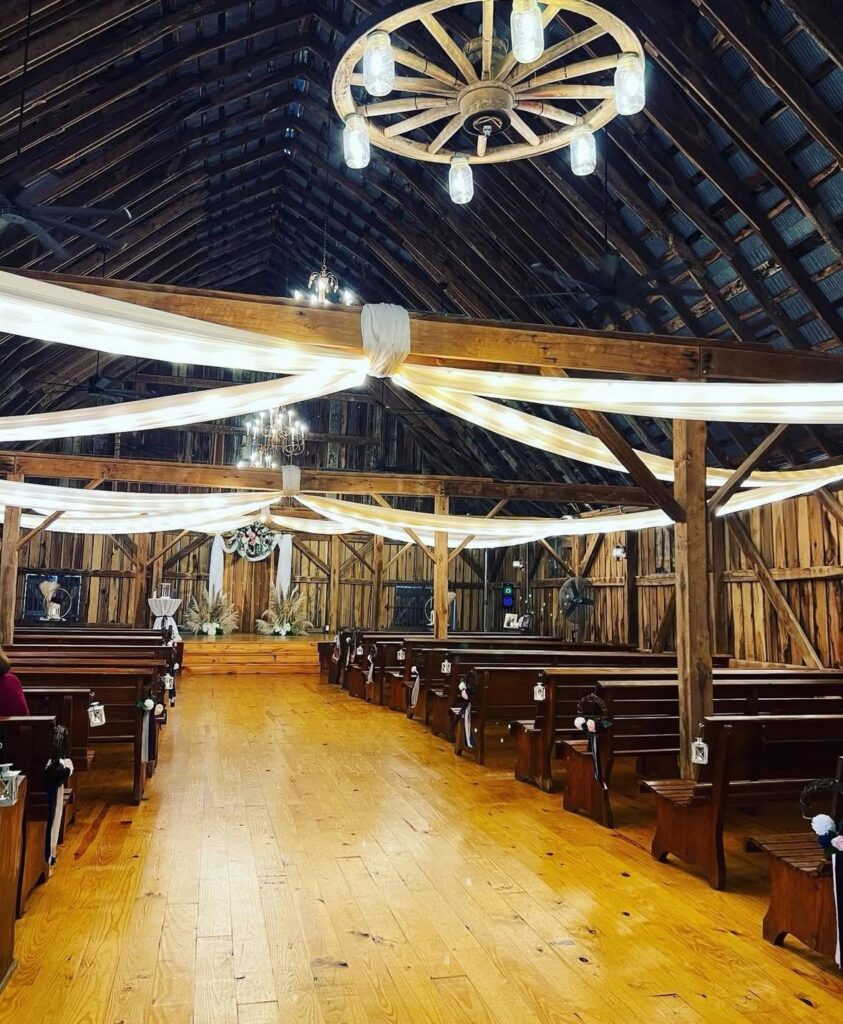 Ceremony hall at Ava's Place barn venue with wooden pews, draped beams and a wagon-wheel chandelier